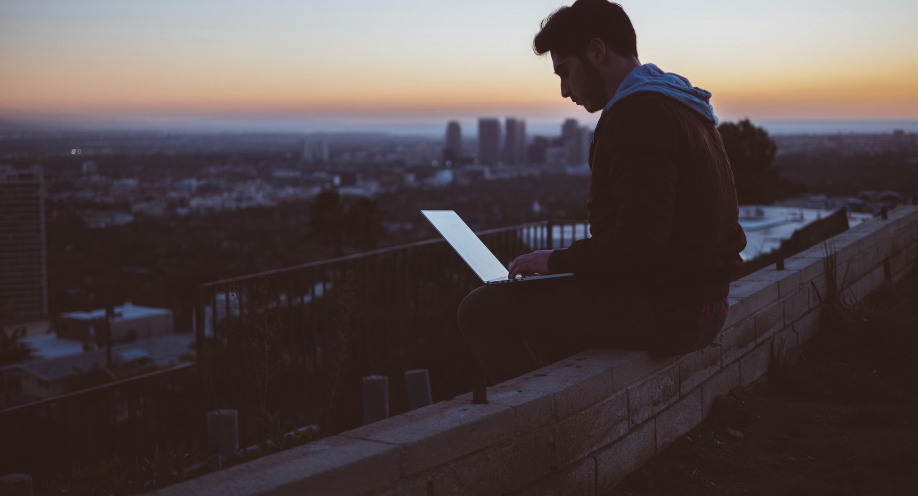 A person working on a laptop on a wall above a city at dusk