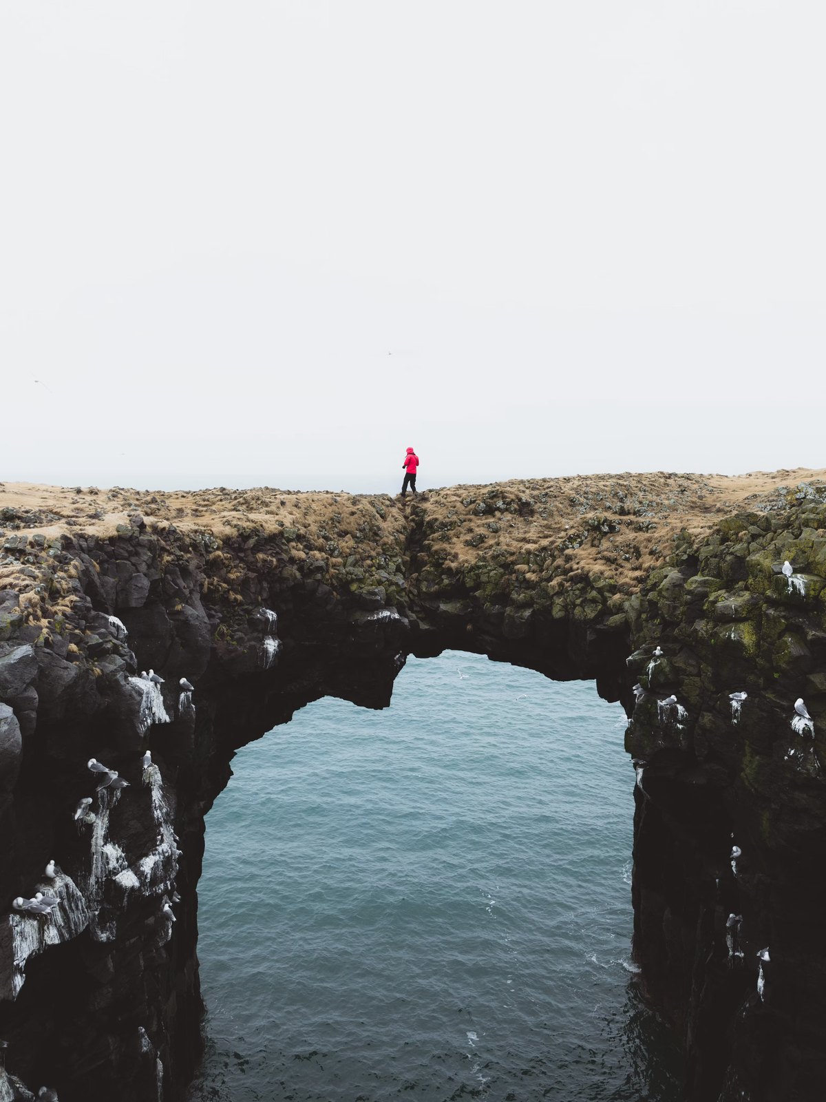 A lone figure standing on a narrow rock arch high above the sea