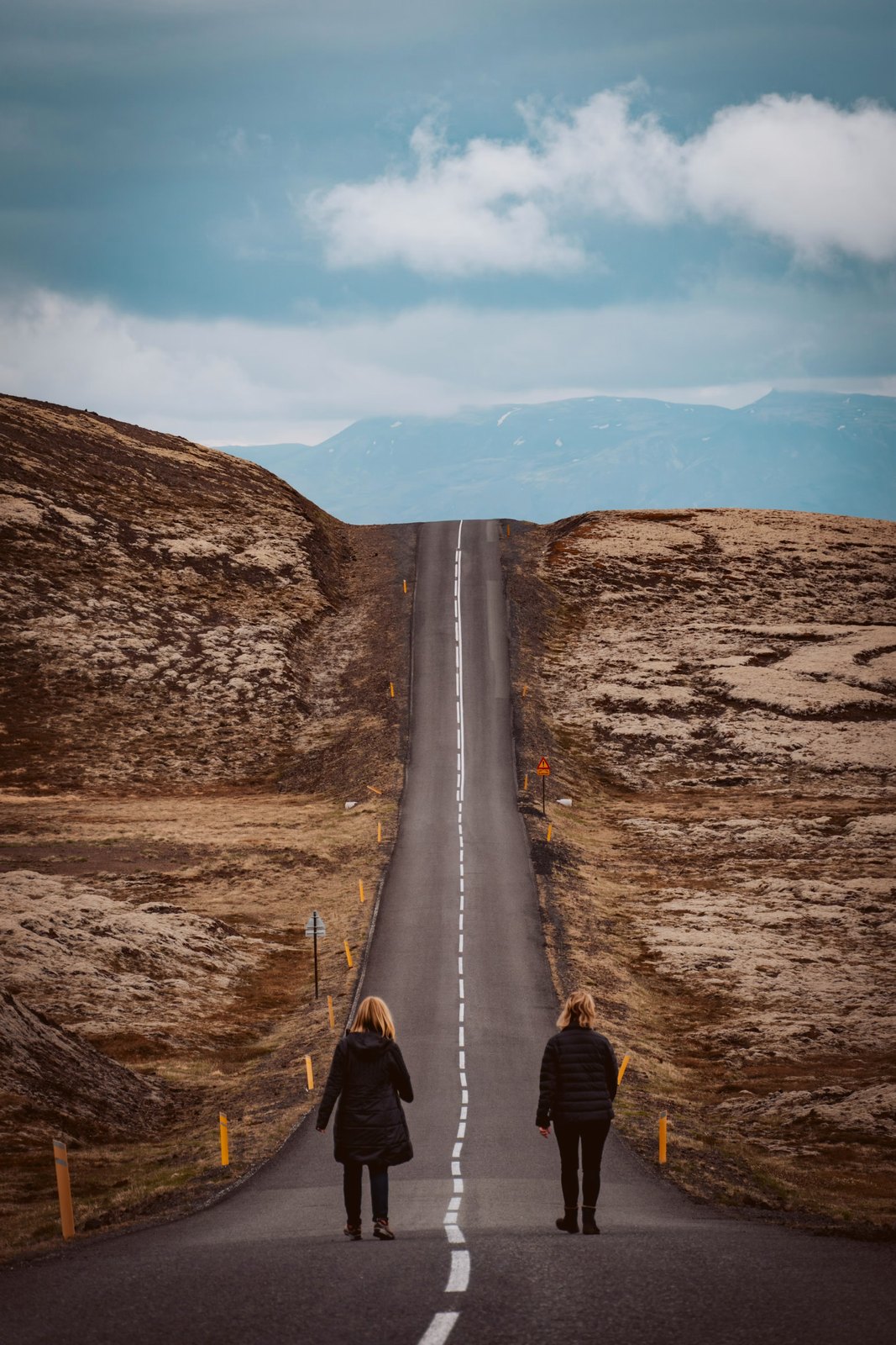 Two people walking a long open road, one slightly ahead of the other