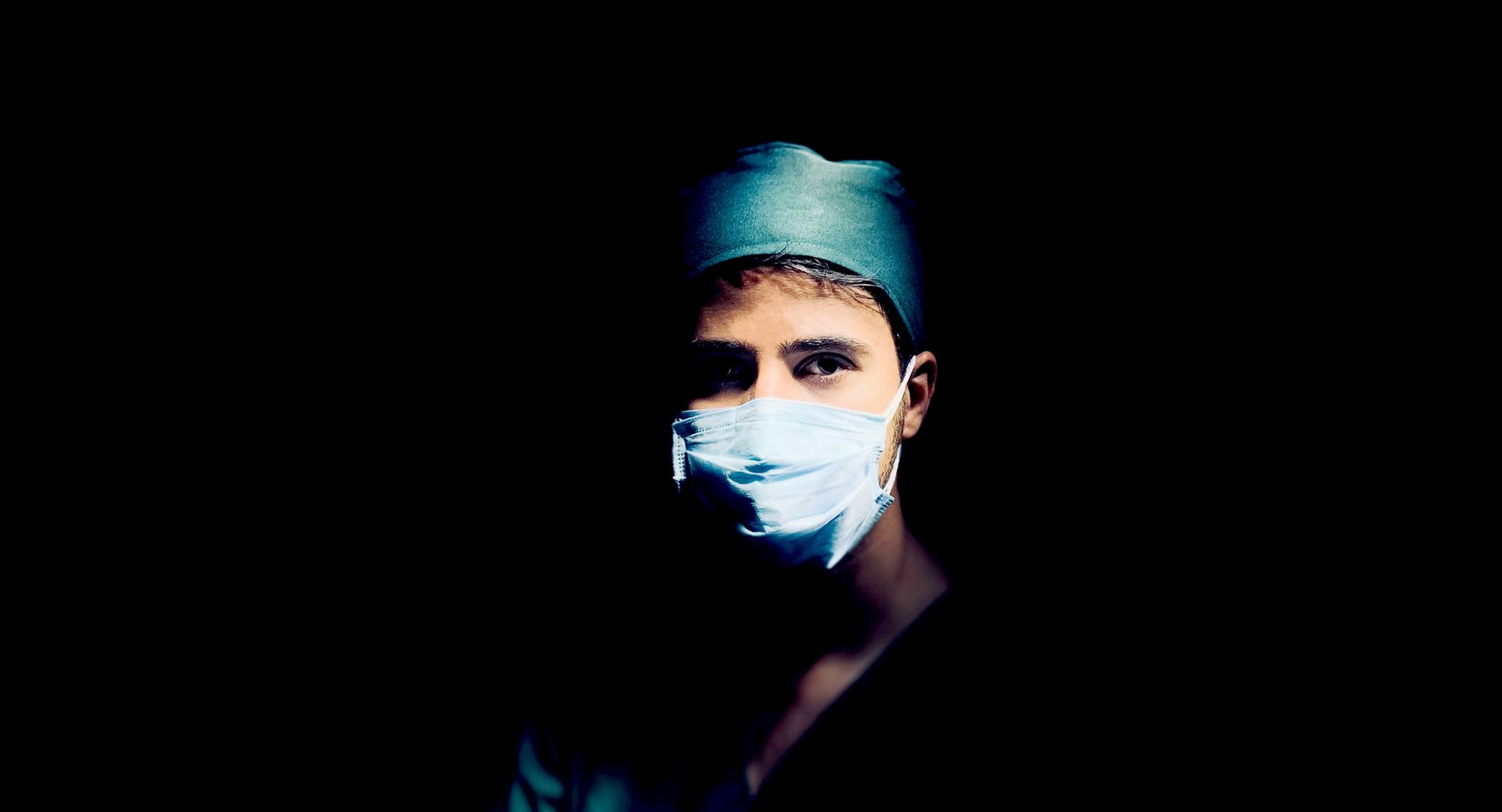 A masked clinician under theatre lights in a darkened room